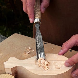 May include: A metal wood gouge with a light-colored wooden handle is being used to carve a shallow, rounded depression into a block of light-colored wood. Small wood shavings are scattered around the carved area on the surface of the block.