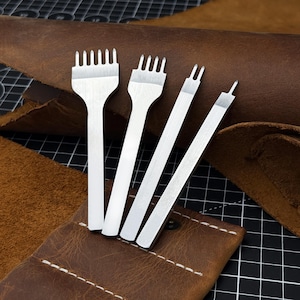 Può includere: Quattro strumenti argentati per la lavorazione della pelle, tra cui ceselli a forcella e uno scanalatore, sono esposti su uno sfondo di pelle marrone e un astuccio in pelle. Gli strumenti sono disposti su una superficie a griglia nera, mostrando il loro design preciso.