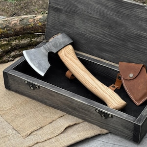 May include: Axe with a wooden handle and a metal blade, presented in a gray wooden box with a black interior. A brown leather sheath is included. The axe is a tool for outdoor activities.