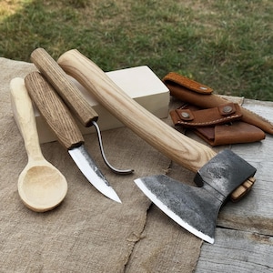 Puede incluir: Un conjunto de herramientas para trabajar la madera, que incluye un hacha pequeña, cuchillos y una cuchara. El hacha tiene un mango de madera y una hoja de metal oscuro. Los cuchillos tienen mangos de madera y hojas de metal. También hay una funda de cuero.