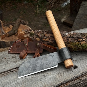 Puede incluir: Una hachuela forjada a mano con una hoja de metal oscuro y texturizado y un mango de madera clara. La hoja presenta un pequeño logotipo estampado. Una funda de cuero marrón con botones a presión color latón y un logotipo a juego está cerca.