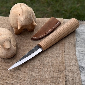Puede incluir: Un cuchillo artesanal con mango de madera y funda de cuero descansa sobre una superficie de arpillera. También hay dos tallas de madera de un elefante y un pato. La hoja del cuchillo es plateada y el mango es de color marrón claro.