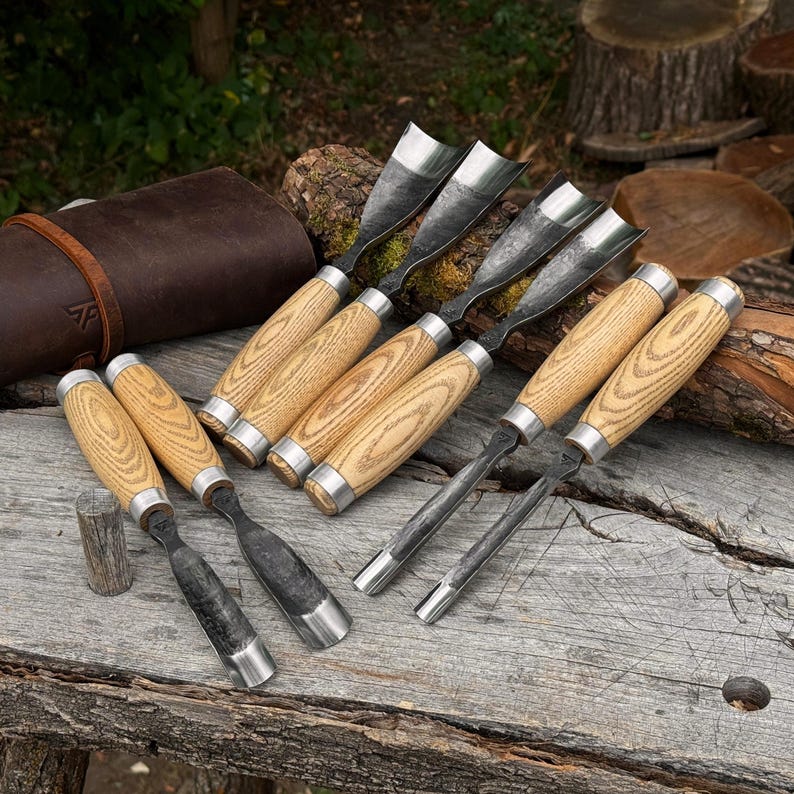 May include: A set of eight wood carving gouges with curved metal blades and light-coloured wooden handles with visible grain patterns. The tools feature silver-toned metal ferrules connecting the blades to the handles, arranged on a rough surface.