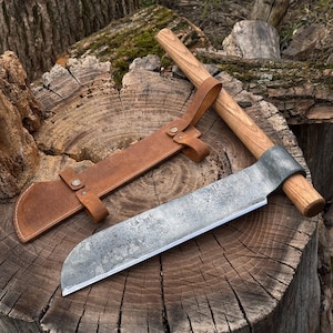 May include: A large, hand-forged cleaver with a wooden handle and a leather sheath. The blade is made of steel and has a textured finish. The sheath is brown leather with a strap and button closure. The cleaver is resting on a wooden surface.