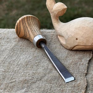 Op de afbeelding: Een houtsnijbeitel met een houten handvat en een metalen lemmet ligt op een getextureerd, beige oppervlak. Op de achtergrond staat een gesneden houten walvis. De beitel heeft een zilveren metalen band.