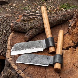 Peut inclure: Deux outils à main dotés de lames en métal gris foncé forgé et de manches en bois brun clair. Chaque lame présente un tranchant droit et un sommet incurvé, avec un petit logo estampillé près du point de fixation du manche sur un tronc.