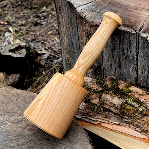 May include: A light-colored wooden mallet with a cylindrical head and a rounded handle. The mallet is made of a light-colored wood, possibly ash or maple, and has a smooth, polished finish. The handle is ergonomically shaped for a comfortable grip. The mallet is resting on a wooden surface.