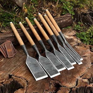 May include: A set of six hand-forged steel chisels with light brown wooden handles, each featuring a wide, flat metal blade and a small stamped logo, arranged in a row alongside a stack of small, rectangular brown leather protective covers.