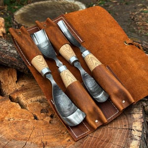 Pu&ograve; includere: Un set di tre sgorbie per intaglio del legno con lame in metallo scuro e testurizzato e manici in legno chiaro, ciascuna dotata di una ghiera in metallo. Gli utensili sono riposti in una custodia avvolgibile in pelle marrone con cuciture bianche.