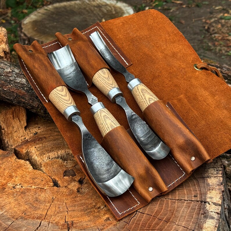 Pu&ograve; includere: Un set di tre sgorbie per intaglio del legno con lame in metallo scuro e testurizzato e manici in legno chiaro, ciascuna dotata di una ghiera in metallo. Gli utensili sono riposti in una custodia avvolgibile in pelle marrone con cuciture bianche.
