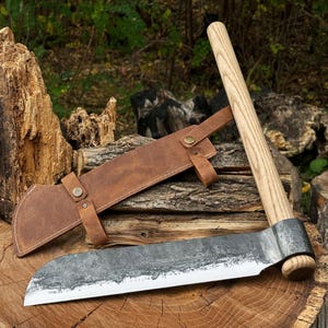 Peut inclure: Une hache forgée à la main avec un manche en bois et un étui en cuir. La tête de la hache a une finition sombre et texturée, et l'étui est marron avec une sangle et une fermeture à bouton. La hache repose sur une surface en bois, avec un arrière-plan extérieur naturel.