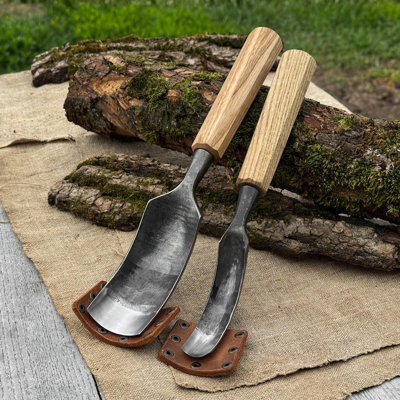 May include: Two hand-forged draw knives with wooden handles and curved blades. The blades are dark gray, and the handles are light brown. Each knife rests on a small, brown leather sheath. The background includes a log and burlap.