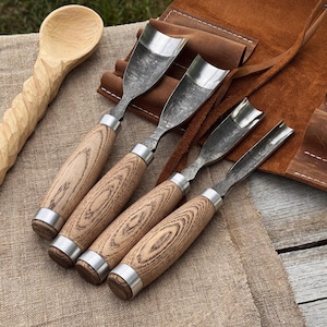 May include: A collection of wood carving tools, including four gouges with wooden handles and metal blades, and a carved wooden spoon. The tools are displayed on a tan surface with a brown leather tool roll in the background.