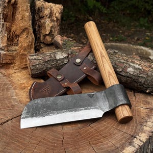 May include: A hand-forged steel hatchet with a straight wooden handle and a dark brown leather sheath. The metal blade features a stamped logo and a sharpened edge, while the leather sheath includes metal snap buttons and a matching logo stamp.