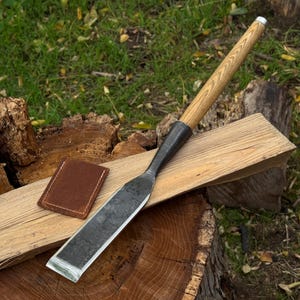 Peut inclure: Un grand ciseau plat avec un manche en bois et une lame en métal, posé sur une planche de bois. Un petit étui en cuir marron est à côté du ciseau. L'outil est pour le travail du bois.