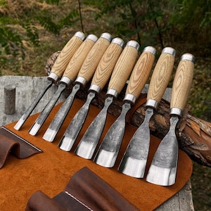 May include: A set of eight wood carving chisels with light-colored wooden handles and silver metal ferrules. Each tool features a forged steel blade with a dark, textured finish and a polished, sharp cutting edge of varying widths.