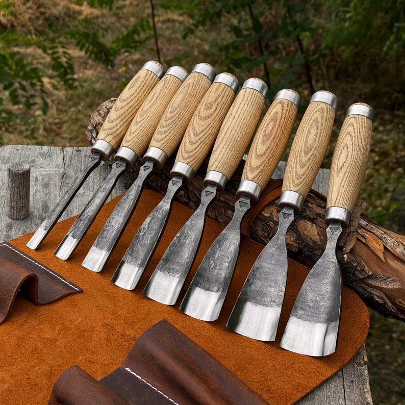 May include: A set of eight wood carving chisels with light-coloured wooden handles and silver metal ferrules. Each tool features a forged steel blade with a dark, textured finish and a polished, sharp cutting edge of varying widths.