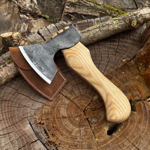 Può includere: Una piccola ascia forgiata a mano con un manico in legno chiaro e una testa in metallo scuro. L'ascia è appoggiata su un ceppo d'albero, con un fodero in pelle marrone che protegge la lama. La testa dell'ascia è larga circa 13 cm.