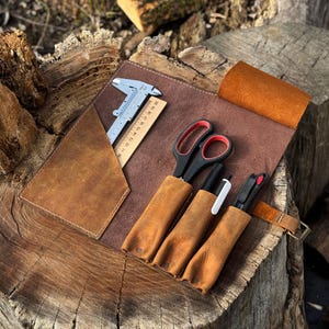 May include: A brown leather tool roll featuring a triangular pocket containing a metal caliper and a wooden ruler marked up to 20 cm. Three additional leather sleeves hold a pair of black scissors with red accents, a black pen, and a utility knife.