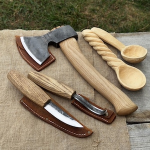 Puede incluir: Una colección de herramientas de carpintería hechas a mano, que incluye un hacha, dos cuchillos con fundas de cuero y dos cucharas de madera. El hacha tiene una cabeza de metal y un mango de madera. Las cucharas tienen detalles tallados.