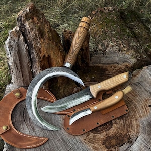 May include: A curved sickle with a wooden handle, a fixed-blade knife with a wooden handle, and a small folding knife with a wooden handle. Each tool is accompanied by a brown leather sheath with metal rivets, displayed on a rough wooden surface.