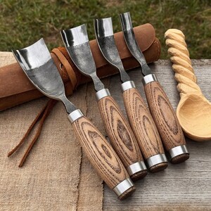 May include: Four wood carving chisels with wooden handles and metal blades. The blades are curved. The handles are light brown with a wood grain pattern. A leather case and a carved wooden spoon are also visible in the image.