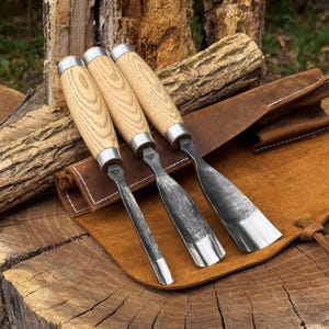 May include: Three wood carving gouges with light-coloured wooden handles and silver metal ferrules. The steel blades feature a dark, textured finish with polished, sharp edges. The tools are arranged side-by-side on a tan leather tool roll.