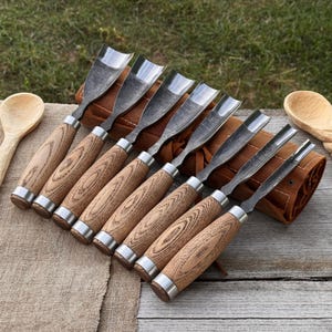 Puede incluir: Un juego de ocho gubias para tallar madera con hojas de acero pulido y mangos de madera. Los mangos tienen un patrón de vetas de madera marrón claro y están acentuados con anillos de metal plateado. Las herramientas están dispuestas sobre un estuche de cuero marrón.
