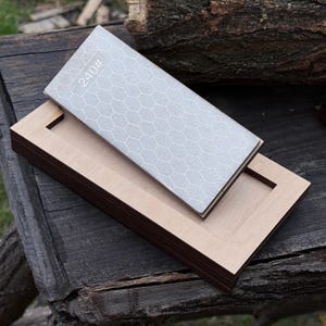 Op de afbeelding: Een grijze slijpsteen met een honingraatpatroon en het nummer "240#" ligt op een houten basis. De basis is gemaakt van lichtgekleurd hout met een donkerbruine rand. De items liggen op een verweerd houten oppervlak.