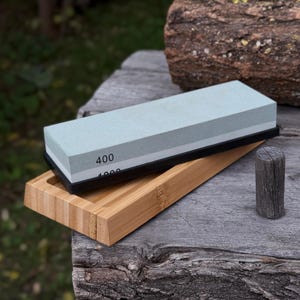 Puede incluir: Un juego de piedras de afilar con una piedra azul claro con las etiquetas "400" y "1000" sobre una base negra, en una bandeja de bambú. Un pequeño cilindro de madera está junto a la bandeja.