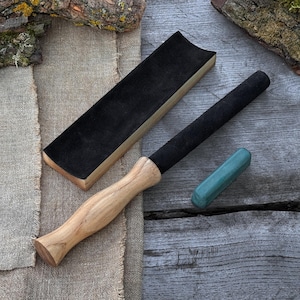 Op de afbeelding: Een slijpstaal met houten handvat en zwart slijpoppervlak, een zwart leren riem en een kleine groene honingsamenstelling. De items worden gepresenteerd op een rustiek houten oppervlak.