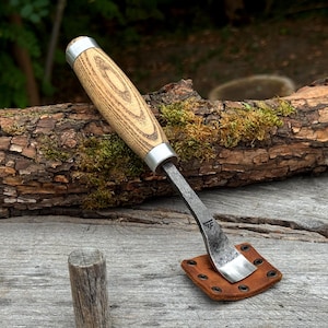 Puede incluir: Una herramienta manual con mango de madera clara, virolas de metal plateado y una hoja de metal gris oscuro curvada. La hoja descansa sobre una pequeña funda de cuero marrón cuadrada con remaches de metal negro en los bordes. Un logo es visible.