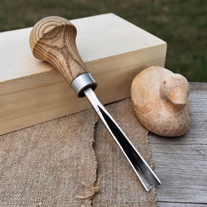 以下が含まれることがあります： 明るい茶色の木製の柄と銀色の口金が付いた、木彫りのノミ。 刃は黒と銀色です。 木彫りのアヒルと木箱も画像にあります。 オブジェクトはジュートの表面にあります。