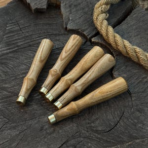 Może przedstawiać: Pięć drewnianych uchwytów do dłuta z mosiężnymi tulejami ułożonych na ciemnej, teksturowanej drewnianej powierzchni. Uchwyty mają jasnobrązowy kolor z widocznym usłojeniem drewna. W tle widoczny jest kawałek liny.