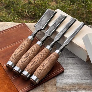 May include: Four wood chisels with wooden handles and metal blades. The handles have a light brown wood grain pattern, and the blades are a dark grey colour. The chisels are arranged on a brown leather surface.