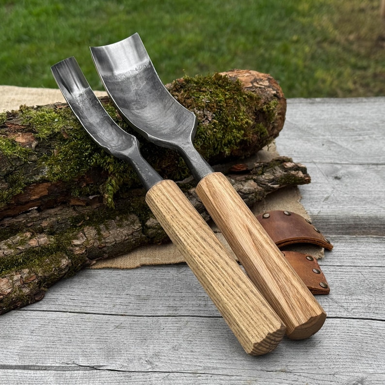 May include: Two wood carving gouges with curved, steel blades and light-colored wooden handles. The tools are displayed on a piece of wood with a leather sheath in the background. The blades have a curved shape, and the handles are cylindrical.