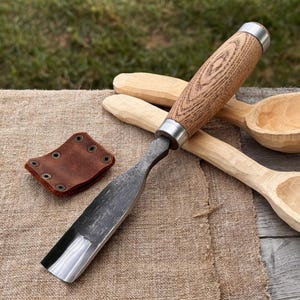 May include: A wood carving gouge with a dark, forged steel blade and a light brown wooden handle with a metal ferrule. Beside the tool lies a small, rectangular brown leather sheath with six metal rivets and two unfinished wooden spoons.
