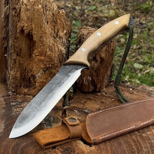 Cuchillo de caza. Hoja fija y afilada. Forjado a mano en acero al carbono 52100 endurecido. Mango ergonómico de fresno. Incluye funda de cuero.