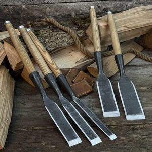 Puede incluir: Un juego de cinco formones para madera con mangos largos de madera clara y hojas de metal forjado oscuro con bordes afilados. Las herramientas están dispuestas en grupo, mostrando su textura metálica y la veta de los mangos de madera.
