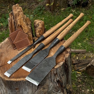 Puede incluir: Un juego de cinceles para carpintería con mangos de madera y hojas de metal, exhibido sobre un tocón de árbol. Los cinceles tienen un acabado de metal oscuro y están dispuestos con algunos posavasos de cuero. El fondo presenta hierba verde y un entorno de madera natural.
