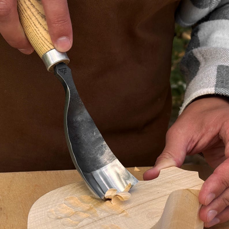 Puede incluir: Una herramienta para tallar madera con una hoja de metal oscuro curvada y un mango de madera clara. La herramienta se muestra en el proceso de tallar una pieza de madera clara, con una peque&ntilde;a viruta de madera visible en la punta.