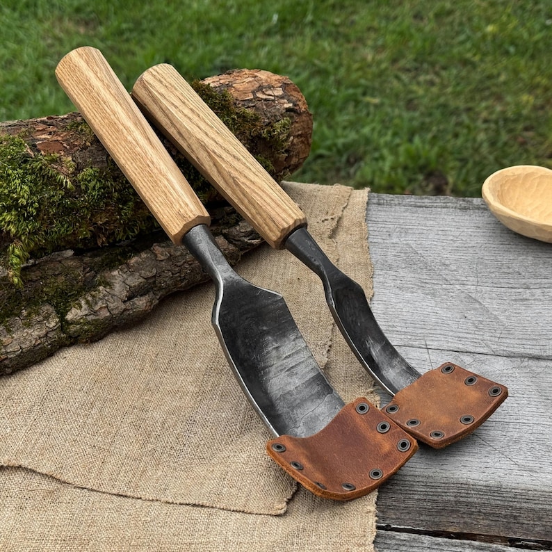 May include: Two hand-forged spoon carving gouges with wooden handles and leather sheaths. The blades are dark metal, and the handles are light-colored wood. The leather sheaths are brown and secured with metal rivets. The tools rest on a wooden surface.