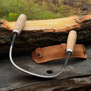 May include: Two curved draw knives with wooden handles and a brown leather sheath. The blades are silver, and the handles have a light wood grain pattern. The sheath has a snap closure. The tools are designed for woodworking.