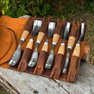 May include: A set of seven wood carving gouges with curved metal blades and light-colored wooden handles, each secured in a brown leather tool roll with individual slots and white stitching, arranged side by side on a weathered wooden surface.