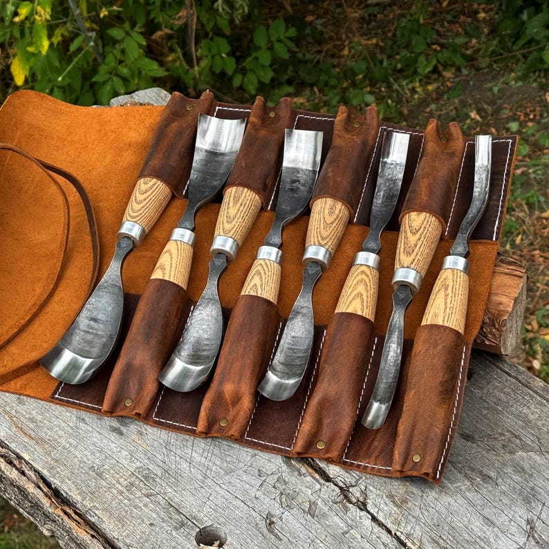 May include: A set of seven wood carving gouges with curved metal blades and light-colored wooden handles, each secured in a brown leather tool roll with individual slots and white stitching, arranged side by side on a weathered wooden surface.