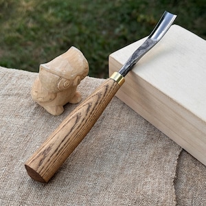 May include: A wood carving gouge with a curved blade and a wooden handle. The handle is made of light brown wood with dark brown grain. A small, carved wooden owl is in the background, along with a light-colored wooden block.