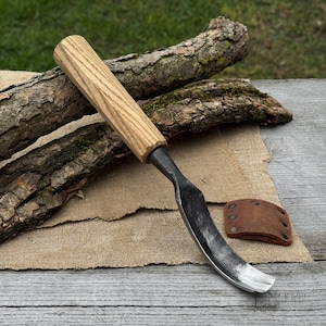 May include: A curved wood carving knife with a light brown wooden handle and a dark metal blade. A small brown leather sheath with rivets is next to the knife. Two pieces of bark are behind the knife.