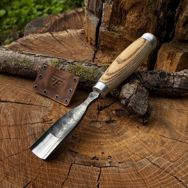 May include: A wood carving gouge with a curved, dark metal blade and a light-coloured, grained wooden handle with silver-toned metal ferrules. Beside the tool lies a small, rectangular brown leather sheath featuring a stamped logo and rivets.
