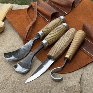 Puede incluir: Un juego de cuatro herramientas para tallar madera con mangos de madera y hojas de metal, expuestas sobre un rollo de herramientas de cuero marrón. Las herramientas incluyen una gubia curva, una hoja recta y un cuchillo de gancho.
