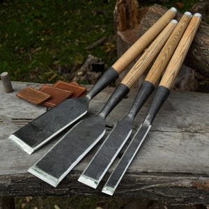 May include: A set of four woodworking chisels with dark, forged metal blades and light-colored wooden handles. The chisels are arranged in a row, showcasing varying blade widths, each featuring a sharp, polished edge and a textured finish.